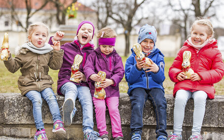 5 Kinder sitzen auf einer Mauer und halten Schokoosterhasen in der Hand auf Schloss Hof.