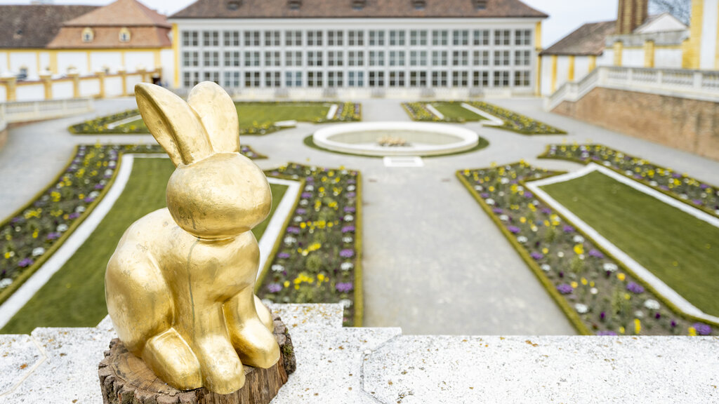 Goldener Hase steht auf einem Geländer, im Hintergrund Barocke Gartenanlage und Orangerie von Schloss Hof.