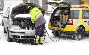 EIn Pannenhelfer steht vor einem Auto mit geöffneter Motorhaube und gibt Starthilfe. Schnee liegt auf der Straße und es schneit.
