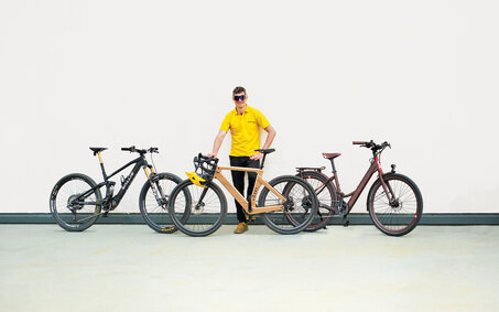 auto touring-Redakteur mit 3 E-Bikes unter 20 kg vor weißer Wand. Das Wallis Project S, das MyEsel E-Gravel und das Kraftstoff E-Razzo Wave.