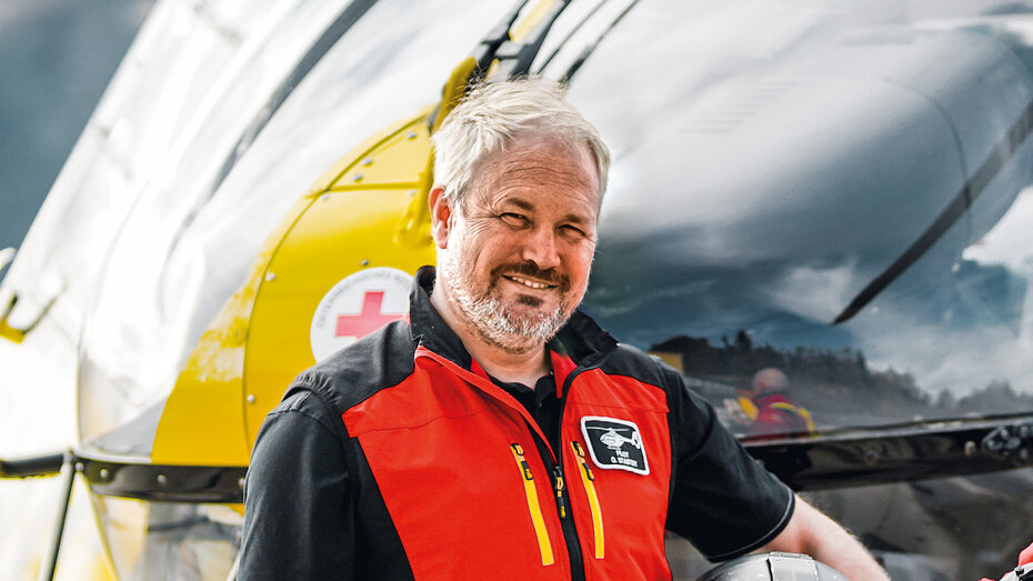 Älterer Hubschrauberpilot von der ÖAMTC Flugrettung. Er trägt eine rote Warnweste, hält seinen Helm und lächelt in die Kamera.