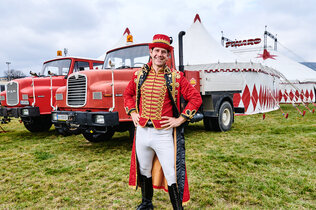 Zirkusdirektor Alexander Schneller steht im Kostüm vor seinen Zugfahrzeugen und dem Zirkuszelt.