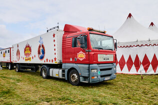 Ein rotes LKW-Zugfahrzeug mit 2 Anhängern des Circus Pikard steht vor dem Zirkuszelt.