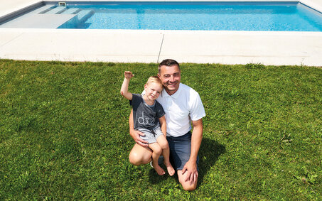 Emil kerngesund und winkend mit seinem Vater im Rasen vor dem Pool.