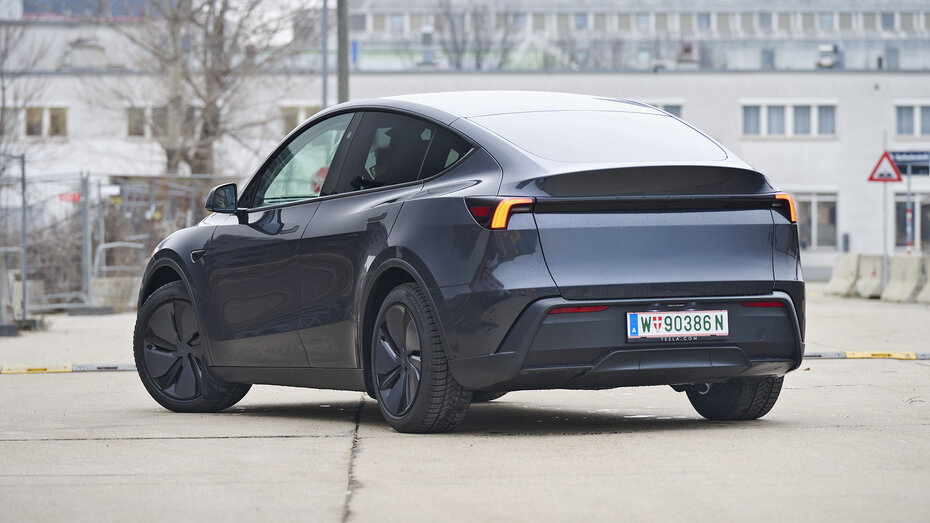Heck eines dunkelgrauen Tesla Model Y, das auf einer Straße vor einem hellgrauen Gebäude steht.