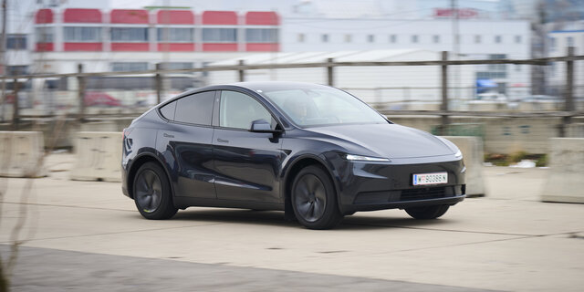 Ein dunkelgrauer Tesla Model Y fährt auf einer Straße vor einem grauen Gebäude.