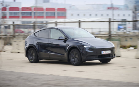 Ein dunkelgrauer Tesla Model Y fährt auf einer Straße vor einem grauen Gebäude.