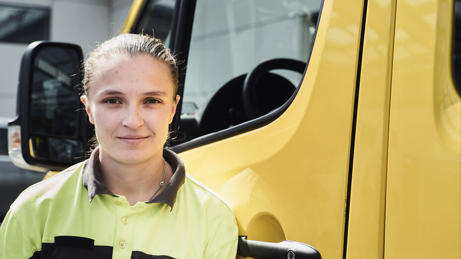 Nahaufnahme: Frau in Arbeitskleidung steht vor einem gelben Auto.