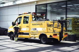 Eine Pannenfahrerin steht vor ihrem kombinierten Abschlepp und Pannenfahrzeug, eine Art Klein-Lkw, vor einem Stützpunkt.