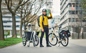 Redakteur mit gelber Jacke und geschulterten Wasserflaschen steht zwischen einem Lastrad und einem Fahrrad mit Gepäcktaschen.