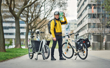 Redakteur mit gelber Jacke und geschulterten Wasserflaschen steht zwischen einem Lastrad und einem Fahrrad mit Gepäcktaschen.