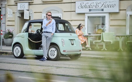 Eine Person in blauem Hemd und grauer Hose lehnt an einem türkisen Microcar, das vor einem Eissalon in der Stadt parkt, und isst Eis.