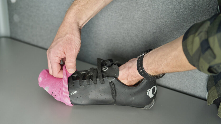 Eine Hand zieht über einen Radschuh einen pinkfarbenenn Socken ziehen. Auch das Bein ist zu sehen. 