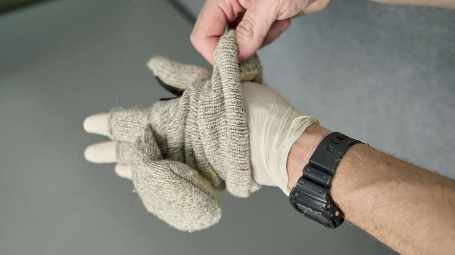 Hände ziehen Handschuhe über Plastik-Einweghandschuhe an. Eine Hand trägt eine Uhr.