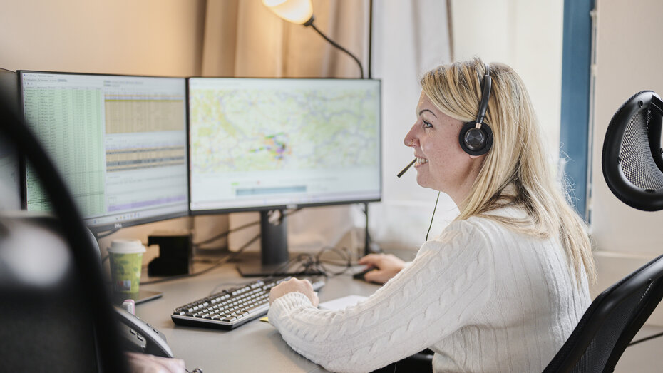 Eine blonde Frau mit Headset zum Telefonieren sitzt vor einem Computer in einem Büro und blickt auf den Bildschirm.