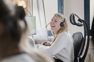 Eine blonde Frau mit Headset zum Telefonieren sitzt vor einem Computer und lacht in einem Büro. Links von ihr ist eine Person im Vordergrund zu sehen. 