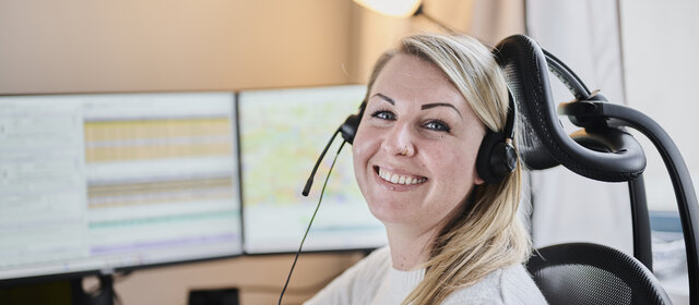 Eine blonde Frau (Victoria Rödleithner) mit Headset zum Telefonieren sitzt vor einem Computer und lacht.
