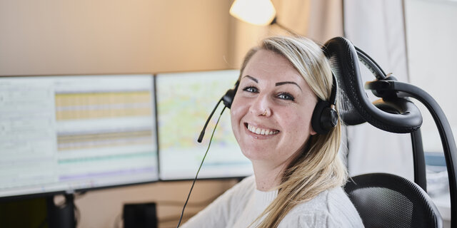 Eine blonde Frau (Victoria Rödleithner) mit Headset zum Telefonieren sitzt vor einem Computer und lacht.