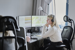 Eine blonde Frau mit Headset zum Telefonieren sitzt vor einem Computer in einem Büro und blickt auf den Bildschirm. Links neben ihr ist das Kopfteil des Sitzes mit ihrer Kollegin zu sehen. 