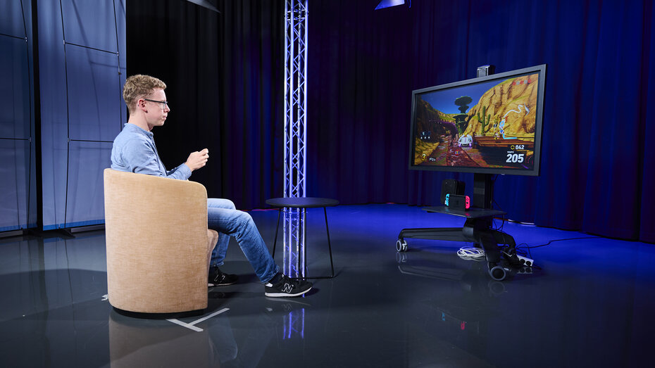 Redakteur Jakob Stantejsky sitzt einem braunen Sessel und spielt mit einem PS5 Controller.