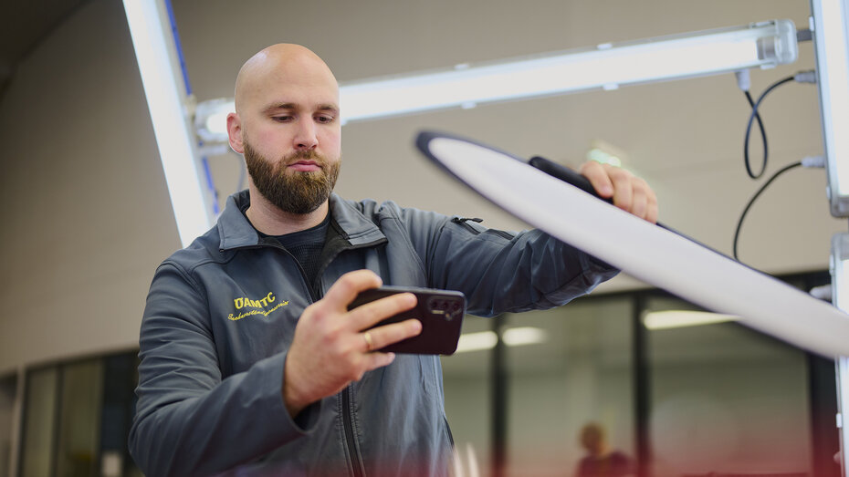 Ein Schadensbegutachter prüft mit dem Dellenspiegel ein Fahrzeug und fotografiert mit dem Handy. 