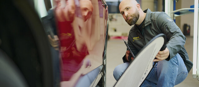 Ein Schadensbegutachter prüft mit einem Dellenspiegel die Tür eines roten Fahrzeugs.