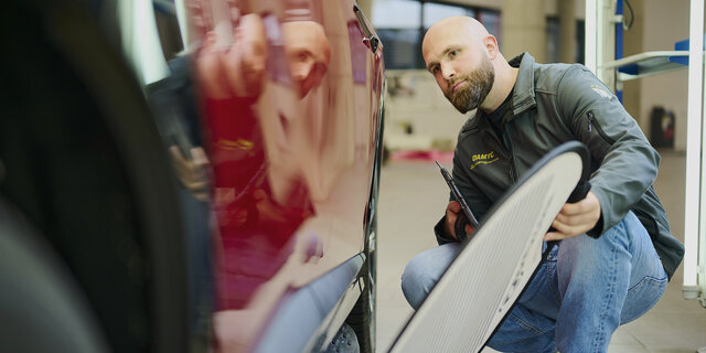 Ein Schadensbegutachter prüft mit einem Dellenspiegel die Tür eines roten Fahrzeugs.