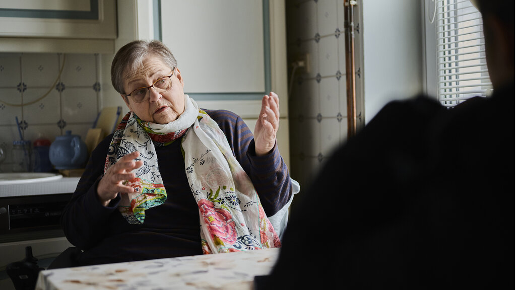 Christine H. erzählt. Sie sitzt an einem Küchentisch. Sie hat kurzes graues Haar und eine Brille.