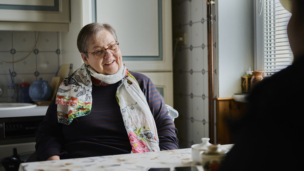 Christine H. erzählt. Sie sitzt an einem Küchentisch. Sie hat kurzes graues Haar und eine Brille.
