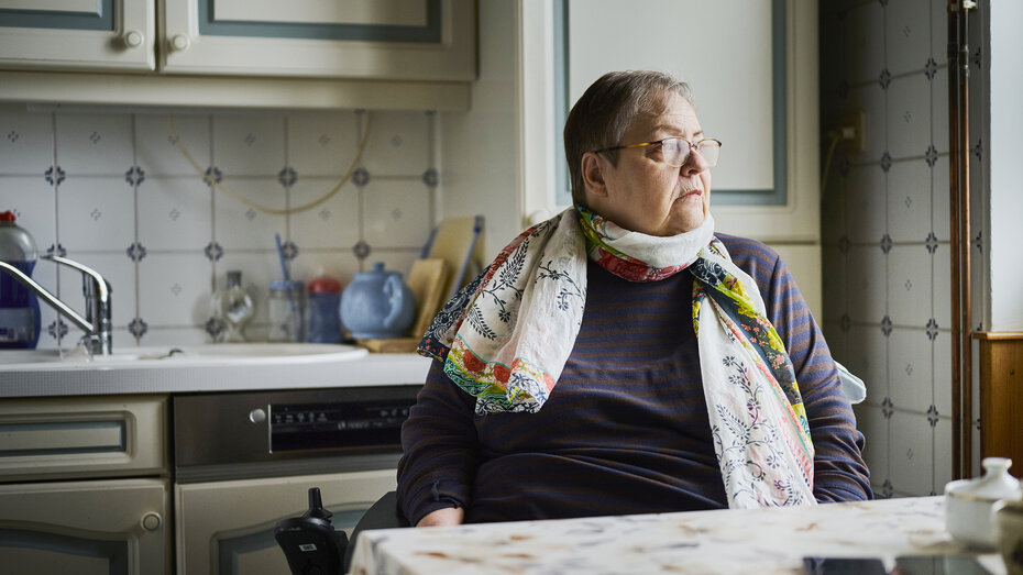 Christine H. schaut aus dem Fenster. Sie sitzt an einem Küchentisch. Sie hat kurzes graues Haar und eine Brille.