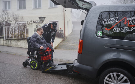 Christine H. fährt in das Taxi. Der Taxifahrer hilft ihr. Sie hat kurzes graues Haar und eine Brille.