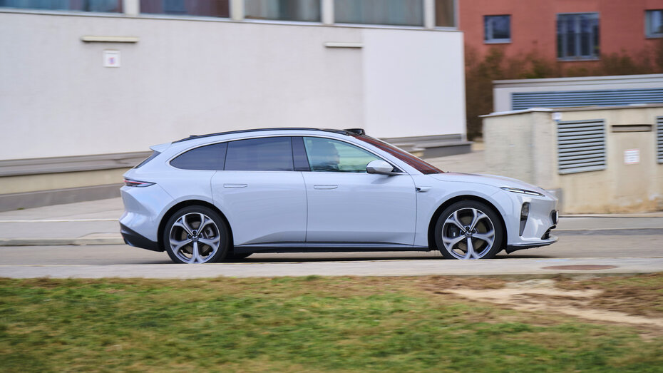 Ein hellblauer Nio ET5 Touring fährt auf einer Straße durch einen urbanen Raum, im Hintergrund befindet sich eine weiße Mauer und ein rotes Gebäude.