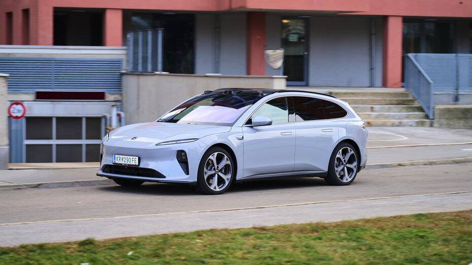 Ein hellblauer Nio ET5 Touring fährt auf einer Straße durch einen urbanen Raum, im Hintergrund befindet sich ein rotes Gebäude.