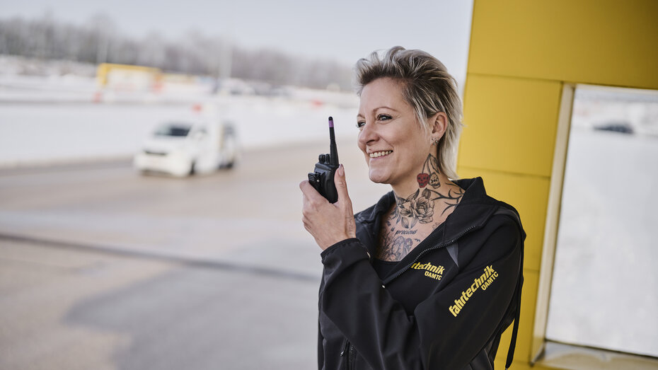 ÖAMTC Fahrtechnik Instruktorin Patricia steht mit Walkie Talkie am Turm in Marchtrenk. Im Hintergrund ist ein Fahrzeug zu sehen. 