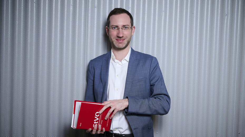 Verkehrsexperte Matthias Nagler steht vor einer grauen Wand mit einem Buch über die Stvo in der Hand. 