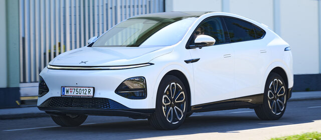 Ein weißer Xpeng G6 fährt auf einer Nebenstraße vor einem Gebäude.