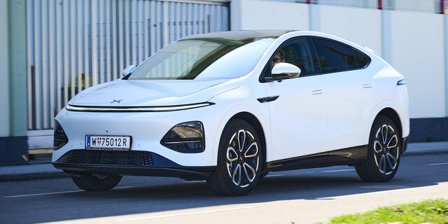 Ein weißer Xpeng G6 fährt auf einer Nebenstraße vor einem Gebäude.