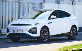 Ein weißer Xpeng G6 fährt auf einer Nebenstraße vor einem Gebäude.