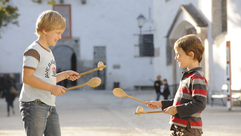 Zwei Buben halten Kochlöffel in der Hand auf einem ist ein Ei platziert. Im Hintergrund ist der Burghof der Hohensalzburg zu sehen.