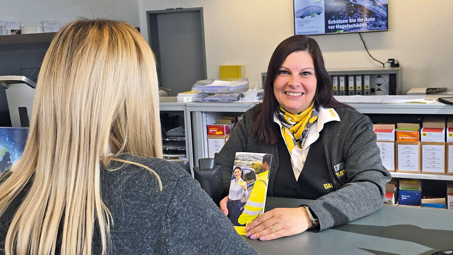 ÖAMTC Stützpunktmitarbeiterin auf ihrem Arbeitsplatz mit einer blonden Kundin. Sie hat braune Haare und lächelt in die Kamera.