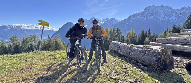 Ein Frau und ein Mann posieren am E-Bike lachend. Im Hintergrund die Berge