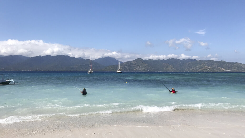 Blick aufs türkise Meer, zwei Fischer stehen im Wasser und ein Segelboot sieht man am Horizont