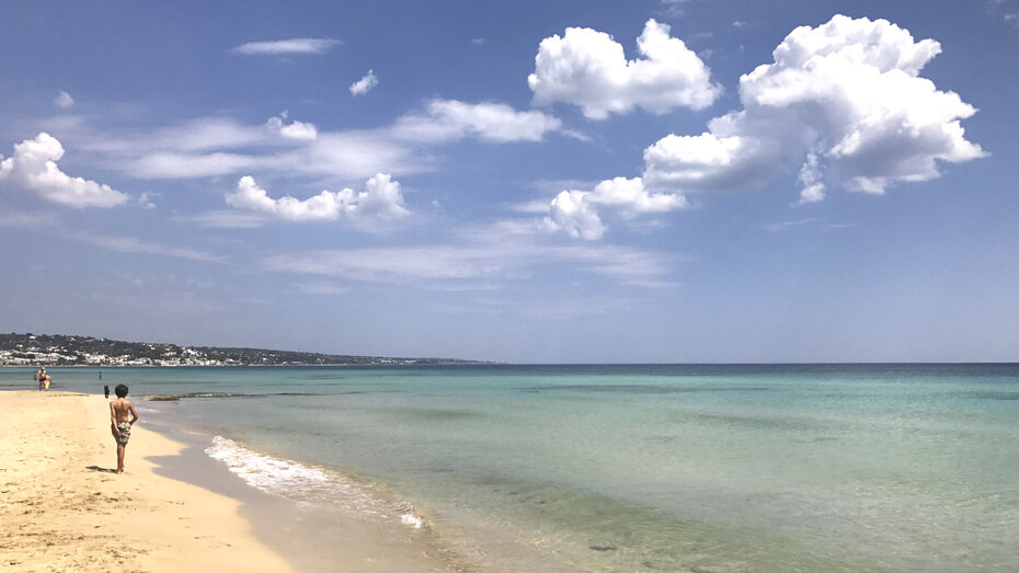 Ein Kind sieht man von hinten und es geht auf einem schönen Sandstrand entlang.