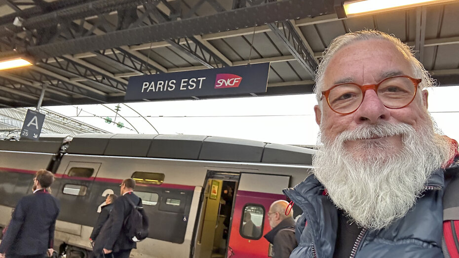 Ein freundlicher Mann mit weißem Bart steht am Bahngleis in Paris, hinter ihm fährt der Zug ein.