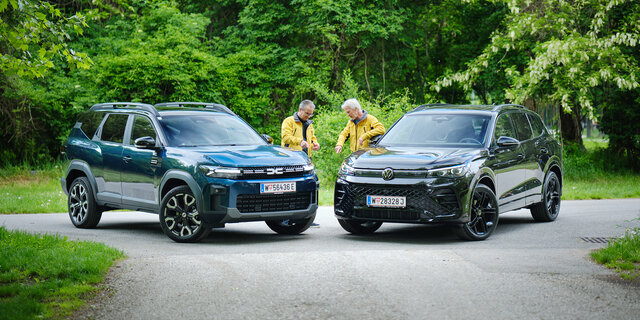Dacia Bigster und VW Tiguan von schräg vorne, dazwischen stehen zwei Testredakteure