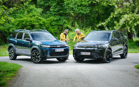 Dacia Bigster und VW Tiguan von schräg vorne, dazwischen stehen zwei Testredakteure