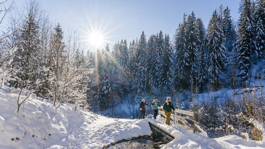 winterwanderer gehen entlang eines Baches mit Wanderstöcken. Die Sonne scheint und der Schnee glitzert.