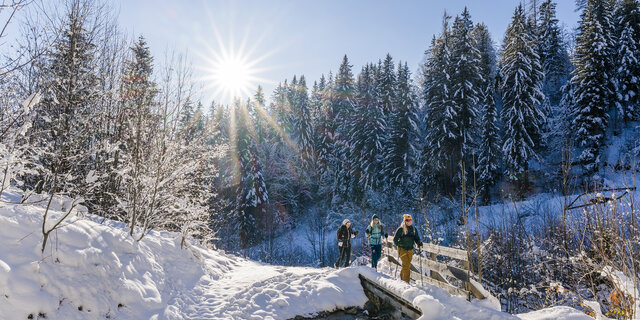 winterwanderer gehen entlang eines Baches mit Wanderstöcken. Die Sonne scheint und der Schnee glitzert.