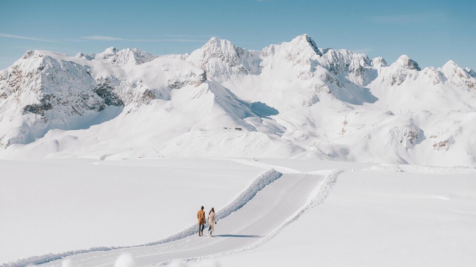Ein Mann und eine Frau spazieren über einen präparierten Weg über ein winterliches Hochplateau, im Hintergrund befinden sich hohe und mit Schnee bedeckte Berge.