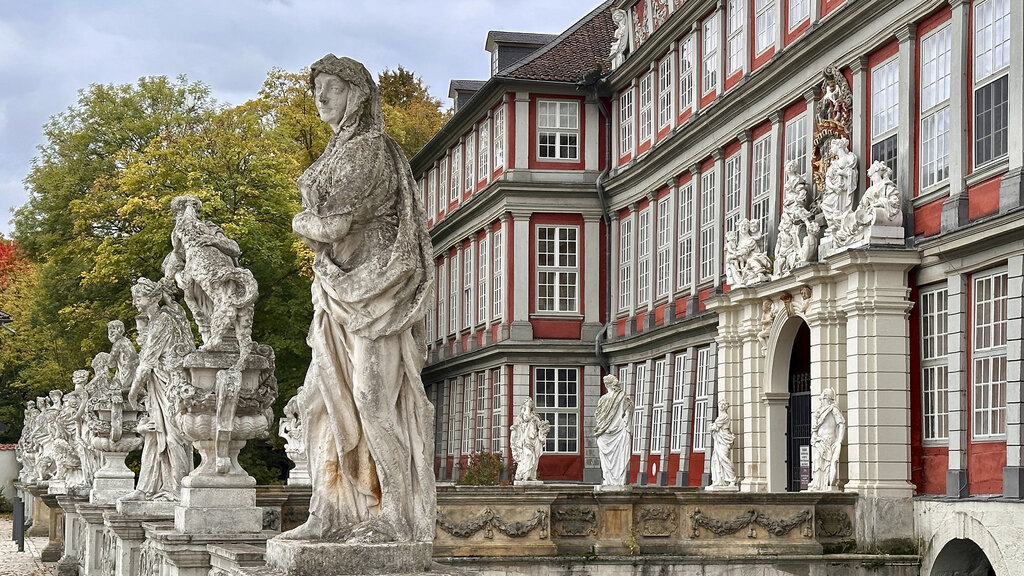 Eine weiße Figurengruppe vor einer Brücke über einen Wassergraben. Am Ende der Brücke ein rotes Barockschloss.